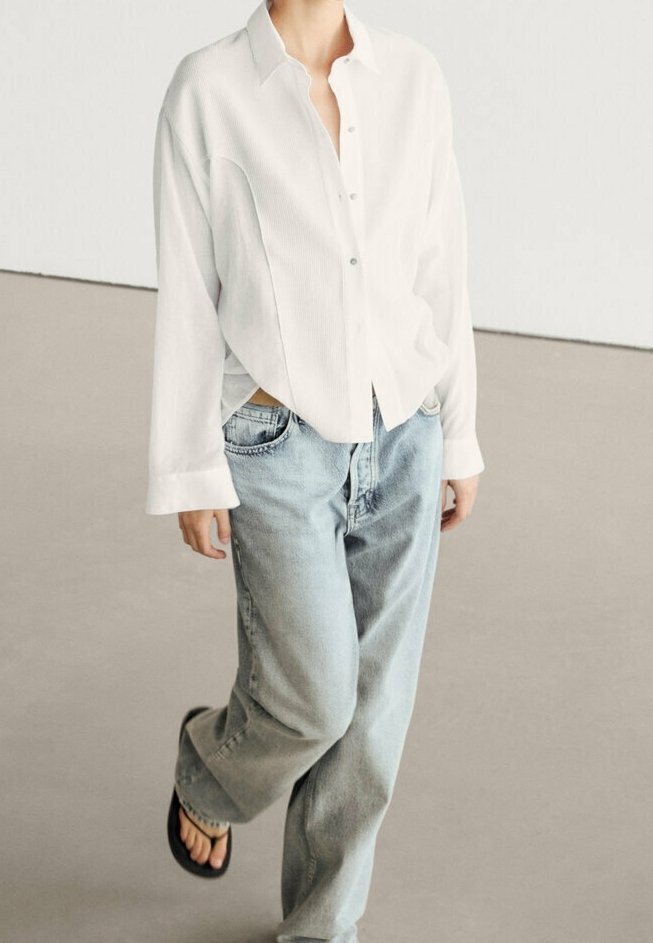 Personne portant une chemise blanche ample boutonnée, un jean large bleu clair et des tongs noires, marchant sur un sol uni.