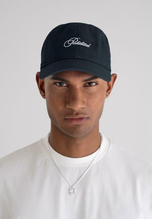 Young man wearing a black cap with "Prohibited" text, a white shirt, and a silver chain necklace against a plain light background.