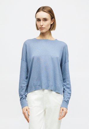 Γυναίκα με κοντά ξανθά μαλλιά, φορώντας ένα ανοιχτό μπλε πουλόβερ με μακριά μανίκια και λευκό παντελόνι, στέκεται μπροστά σε απλό λευκό φόντο.