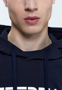Felpa blu navy con tessuto strutturato, dotata di cordoncino, colletto aderente e dettagli del logo bianco audace sul petto.