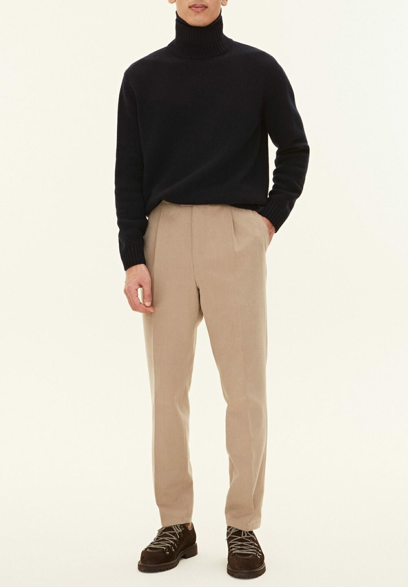 Maglione a collo alto navy, pantaloni beige con pieghe e scarpe marroni stringate con suole testurizzate. Design semplice e palette di colori neutri.