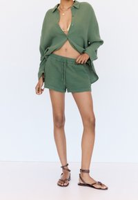 Zielona, teksturowana koszula zapinana na guziki, częściowo rozpięta, noszona na dopasowanych szortach z elastycznym pasem. Brązowe sandały na paski z metalowymi ozdobami.