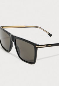 Čierne obdĺžnikové slnečné okuliare s tmavými šošovkami, zlatými akcentami na pántoch a ramenách, a logom „BOSS“ na spánku.