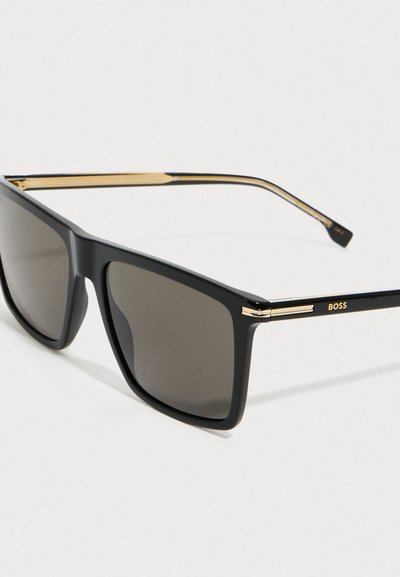 Lunettes de soleil noires rectangulaires avec des verres sombres, accents dorés sur les charnières et les branches, et logo "BOSS" sur la branche.