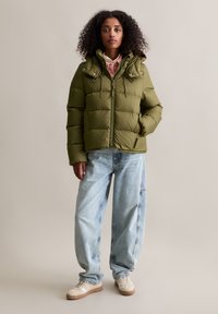 Oliwkowa kurtka puchowa z kapturem, z zamkiem błyskawicznym i przednimi kieszeniami. Nosi się na jasnoniebieskie, luźne dżinsy i kremowe sneakersy.