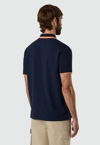 Hombre con cabello corto y castaño, vestido con una camisa polo azul marino con ribetes naranja y blanco, y pantalones cortos cargo beige, de espaldas sobre un fondo liso.