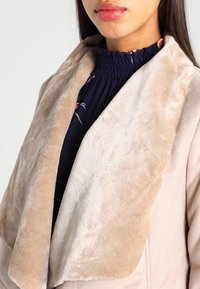 Béžový límec z umělé kožešiny na světle béžovém kabátu s jemnou texturou, detail naznačující nařasený výstřih s tmavými květinovými akcenty pod ním.