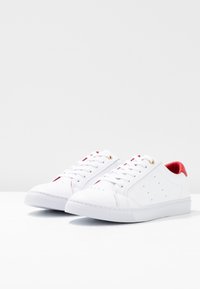 Par de zapatillas blancas de caña baja con forro interior rojo, cordones blancos y pequeños detalles perforados en los laterales.