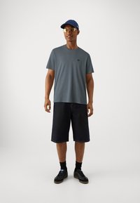 Pelēks T-krekls, tumši džinsu šorti, melni sporta apavi, zila cepure un saulesbrilles. Vienkāršs dizains, īsas piedurknes un brīvs piegriezums ar minimālu zīmola atpazīstamību.