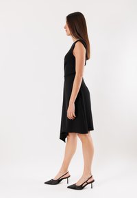 Abito nero senza maniche con orlo asimmetrico, texture liscia e vita aderente, abbinato a scarpe con tacco a stiletto nere e décolleté.