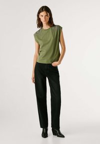 Top sin mangas verde oliva con escote redondeado y adornos plateados en los hombros, combinado con jeans negros de pierna recta y botas tobilleras.