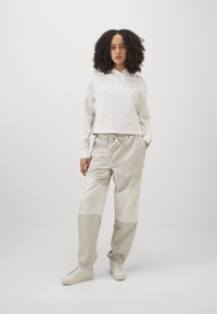 Hanorac scurt alb cu șnur, asortat cu pantaloni de trening gri deschis cu detalii panel. Modelul poartă adidași albi.