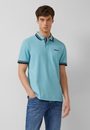 Junger Mann trägt ein hellblaues Poloshirt mit dunkelblauer Umrandung und Jeans, steht vor einem einfarbigen grauen Hintergrund.