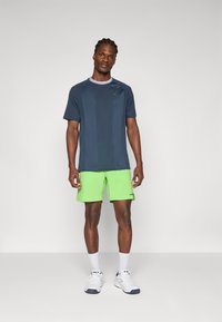Camiseta deportiva azul marino de manga corta y cuello redondo combinada con pantalones cortos de color verde brillante. Zapatillas blancas y calcetines hasta el tobillo completan el conjunto.