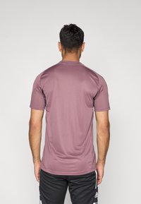 Αθλητική μπλούζα με κοντά μανίκια σε απαλό μοβ χρώμα, κατασκευασμένη από απαλό, ελαφρύ ύφασμα με αντίθετες μαύρες λεπτομέρειες στους ώμους.