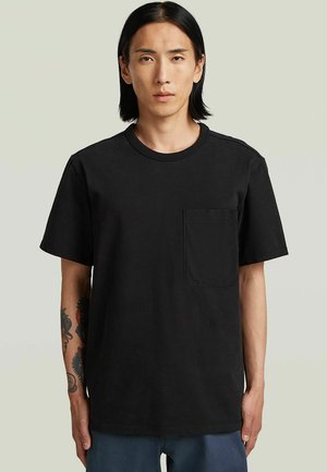 Un homme aux cheveux noirs mi-longs porte un t-shirt noir uni avec une poche poitrine, debout devant un fond clair neutre.