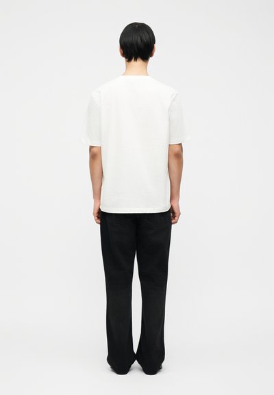 Άτομο με κοντά μαύρα μαλλιά, που στέκεται με την πλάτη προς τα εμάς, φορώντας ένα απλό λευκό κοντομάνικο πουκάμισο και μαύρο παντελόνι ίσιας γραμμής, με φόντο το λευκό.