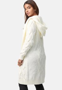 Cardigan tricoté blanc avec une capuche, présentant un design à torsades et des poignets côtelés. Texture douce et longueur longue, adapté pour le superposition.