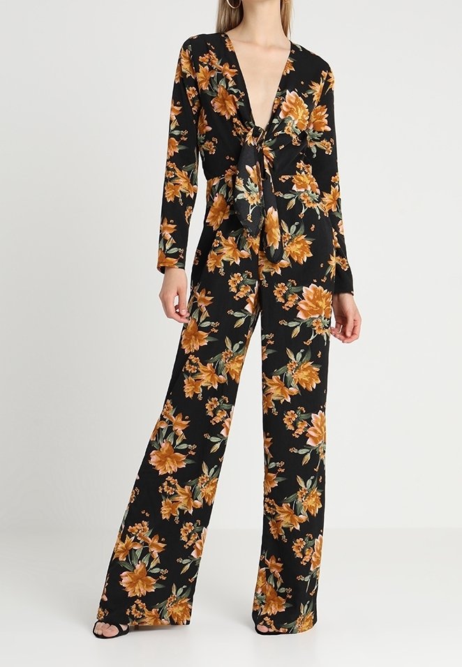Femme portant une combinaison noire à fleurs avec manches longues, jambes larges et un nœud noué sur le devant, debout devant un fond clair uni.