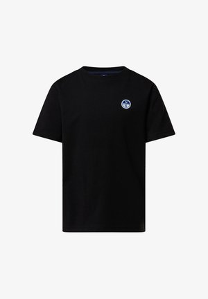 Garçon debout, une main dans la poche, portant un t-shirt noir et un pantalon, tenant une veste noire, et des baskets blanches sur un fond uni.