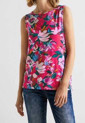 Mouwloos felroze topje met grote kleurrijke bloemenprint, gedragen met een blauwe spijkerbroek door een persoon die tegen een effen achtergrond staat.