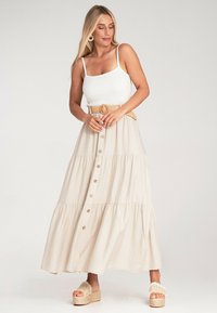 Beige Maxikleid mit gestuftem Design, mit knopflosem Vorderteil und breitem Bund. Modell trägt ein weißes, geripptes Tanktop und beige Sandalen.
