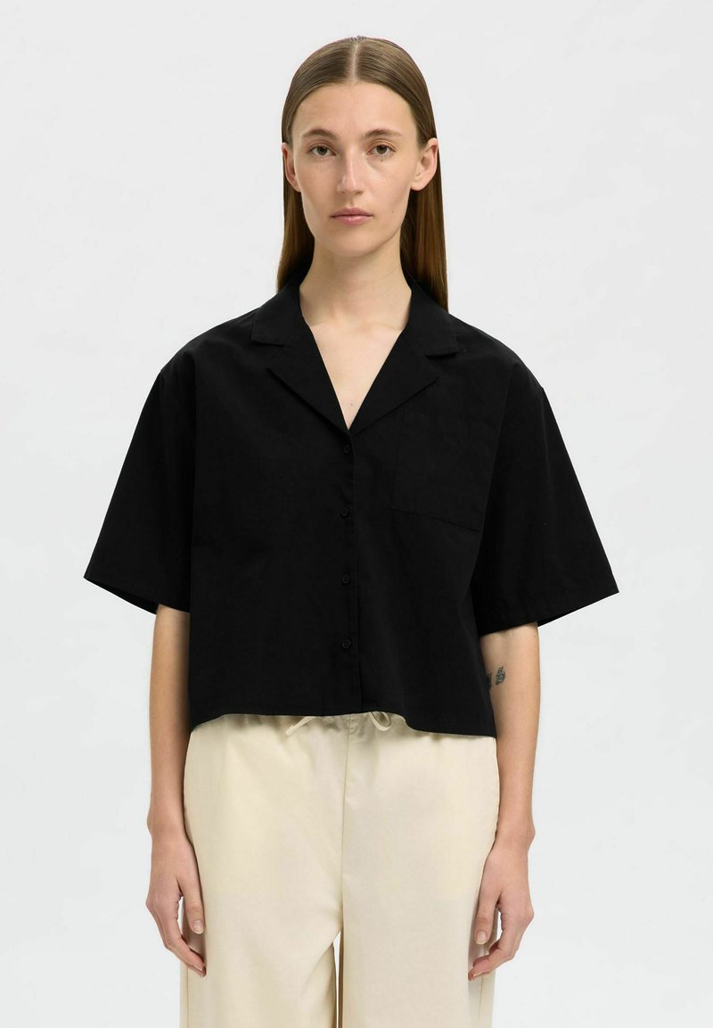 Schwarzes, cropped Button-up-Shirt mit kurzen Ärmeln, flachem Kragen und einer Brusttasche, aus einem glatten Stoff. Getragen mit beigen Hosen.