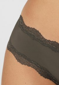 Biancheria intima verde oliva scuro con una texture liscia, caratterizzata da un bordo di pizzo smerlato lungo la cintura per un dettaglio aggiuntivo.