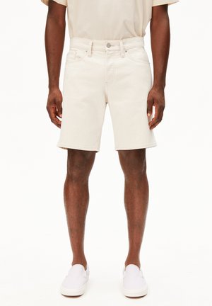 Männliches Model trägt elfenbeinfarbene Jeansshorts, beiges T-Shirt und weiße Slipper, steht vor einem schlichten weißen Hintergrund.