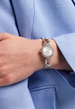 Montre en métal argenté avec un cadran rond, un fond blanc, trois petits points comme indicateurs d'heure, et un bracelet maillon avec des maillons lisses et texturés.