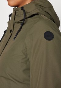 Armygrøn udendørsjakke med hætte, med sorte snore, trykknaplukning og et rundt logo-patch på skulderen.