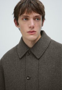 Cappotto di lana marrone con colletto, chiusura a bottoni singola e due bottoni scuri. Il tessuto sembra morbido con una finitura texture.