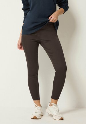 Person iført mørkebrune tætsiddende bukser, en mørk langærmet top og hvide sneakers, der står mod en ensfarvet baggrund.