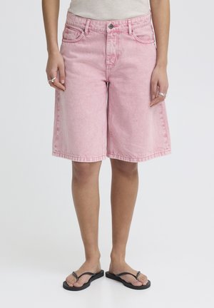 Person iført løse, knælange pink denimshorts og sorte flipflops, stående mod en ensfarvet hvid baggrund.