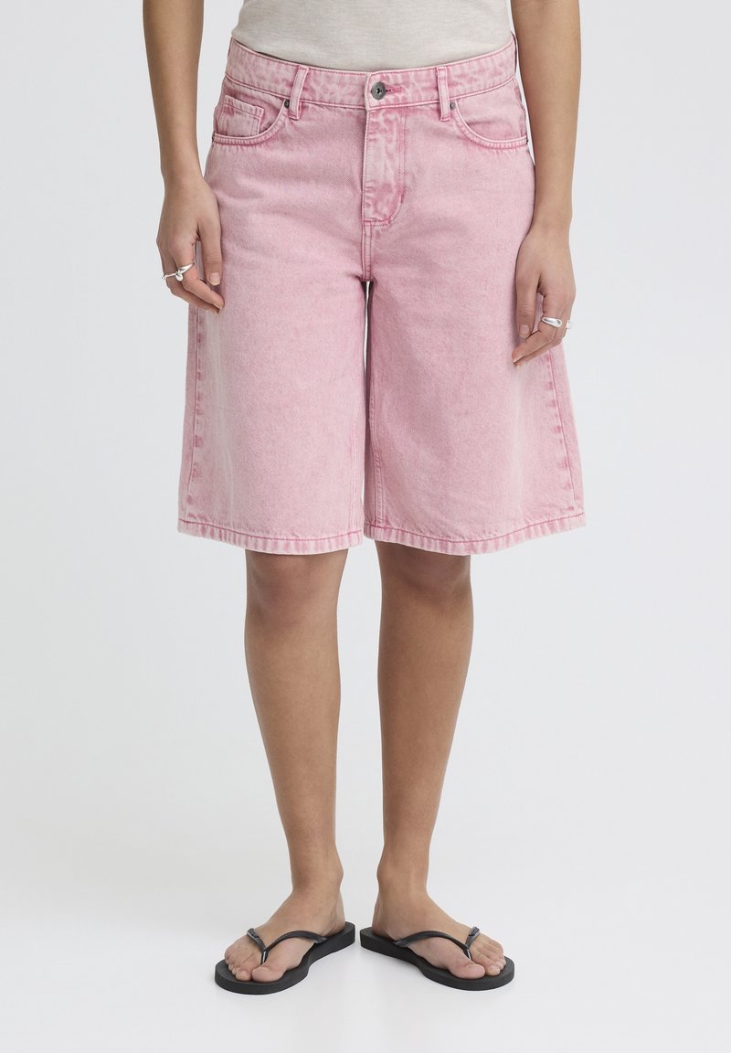 Personne portant un short en denim rose ample, arrivant aux genoux, et des tongs noires, debout devant un fond blanc uni.