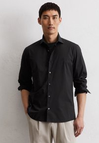 Camicia nera a maniche lunghe con bottoni e colletto, caratterizzata da un logo discreto, una tasca sul petto e maniche arrotolate. Texture liscia.