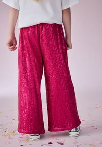 Pantaloni larghi di paillettes fucsia con una texture morbida, dotati di una cintura elastica, indossati con sneakers argento. Sfondo chiaro.