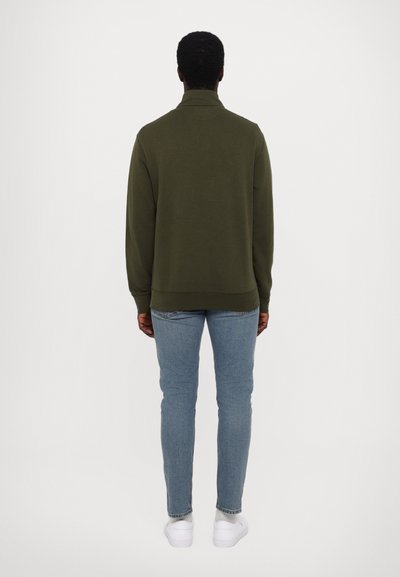 Olivengrøn sweatshirt med høj krave, parret med lyseblå slim-fit jeans og hvide sneakers, set fra bagsiden.