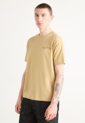 Camiseta de algodón marrón claro con cuello redondo y mangas cortas. Presenta un pequeño logo de la marca impreso en el lado izquierdo del pecho. Pantalones de mezclilla negros.