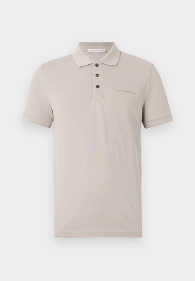 Vaaleanbeige polo-paita, joka on valmistettu pehmeästä kankaasta. Siinä on kaulus, kolme nappia edessä sekä huomaamaton brändäys vasemmalla puolella. Lyhyet hihat.