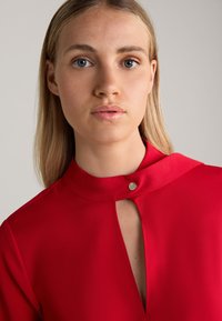 Rode blouse met een hoge kraag en een sleutelgatopening aan de voorkant, met een zilveren knoop accent bij de hals. Soepele stoftextuur.