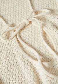 Haut en tricot de couleur crème avec un motif texturé et des liens ajustables. Le tissu a une sensation douce avec des détails de points ouverts.