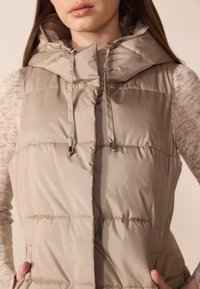 Beige pufferväst med hög krage och dragskohette. Har horisontell sömnad och sidofickor. Materialet verkar mjukt och glänsande.