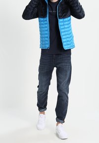 Sininen ja musta pehmustettu takki, jossa on pentukuvio, päällä tummanlaiset paita, yhdistetty sinisiin farkkuihin ja valkoisiin tennareihin.