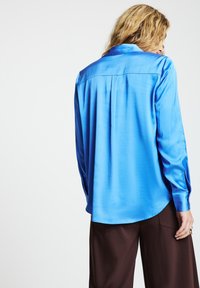 Person with long blonde hair wearing a bright blue satin long-sleeve shirt and dark brown pants, shown from the back against a plain background.