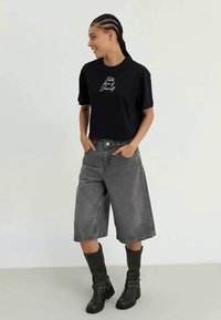 Svart avkortet t-skjorte med fargerik brodert tekst, kombinert med grå denim culottes og høye svarte støvler med spennedetaljer.