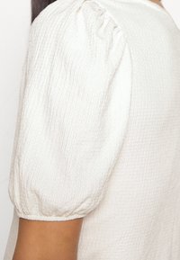 Top de tela blanca texturizada con mangas cortas abullonadas y detalles fruncidos, que exhibe una superficie suave y ligeramente arrugada.