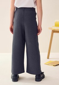 Pantalons gris à jambes larges avec une texture lisse. Présentent une taille haute et aucun matériel visible. Associés à des chaussures noires pour le contraste.