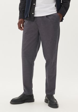 Grau gemusterte Hose mit lockerem Schnitt, ausgestattet mit vorderen Falten und Seitentaschen, kombiniert mit schwarzen Slippern.