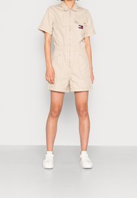Béžový overal s krátkým rukávem a předním zipem, elastickým pasem, dvěma předními kapsami a nášivkou na hrudi. V kombinaci s bíl�ými teniskami.